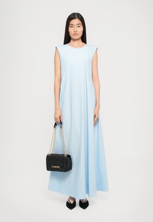 Frau in einem ärmellosen, hellblauen Maxikleid, die eine schwarze, gesteppten Handtasche mit goldenen Kettenriemen hält und schwarze, spitz zulaufende Schuhe trägt.