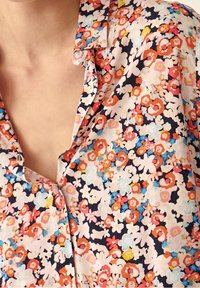 Närbild av en blommönstrad skjorta med orange, vita, rosa och blå blommor mot en mörk bakgrund, delvis uppknäppt vid kragen.