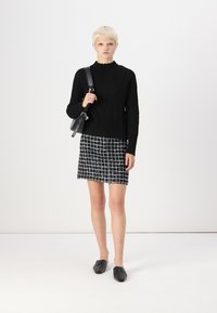 Fekete cable-knit pulóver, kockás fekete-fehér szoknya és lapos fekete cipők. A modell egy fekete táskát tart, amelynek letisztult dizájnja van.