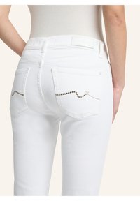 Weiße Jeans aus Denim mit hinteren Taschen, verziert mit einer gewellten Linie aus kleinen Metallic-Nieten. Glatte Textur und figurbetontes Design.