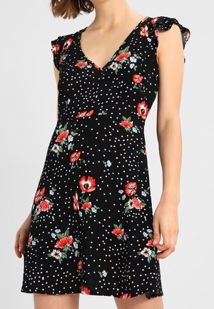 Kvinde iført en sort kjole med hvide polkaprikker og røde blomsterprint, med V-hals og korte flæseærmer.