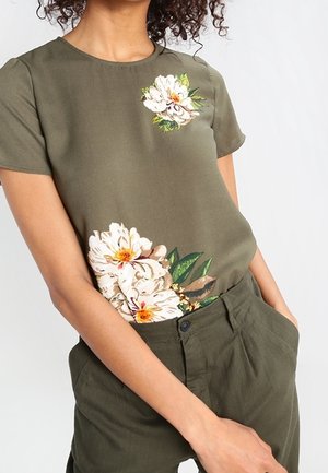 Frau trägt olivgrünes Kurzarmoberteil mit weißen Blumenmustern an Brust und Taille, kombiniert mit passenden olivgrünen Hosen.