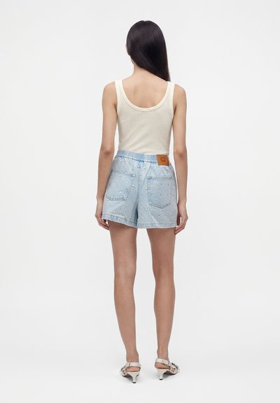 Femme aux longs cheveux foncés portant un haut blanc sans manches, un short en jean taille haute bleu clair avec des clous en forme de perles, et des sandales blanches à talons, debout.