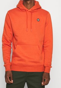 Orange hoodie med en känguruficka, dragsko i huvan och svart logotyp patch på bröstet. Mjuk tyg med en avslappnad passform.
