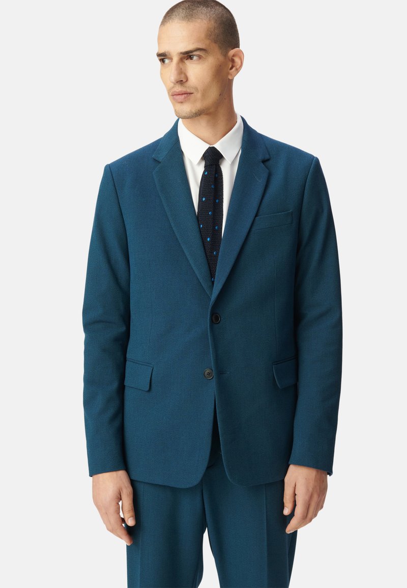 Blazer teal realizzato in tessuto testurizzato, con due tasche frontali, rever a punta e chiusura con un solo bottone. Accompagnato da una camicia bianca e una cravatta a pois.