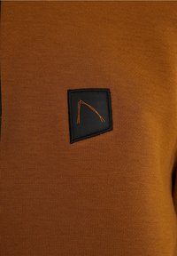 Tissu marron avec un petit patch noir présentant un motif de lignes abstraites orange cousu au centre.