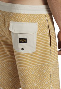 Pantaloni da nuoto gialli con sezioni a righe verticali e a rombi. Presentano una tasca posteriore bianca con una patch logo nera e chiusura a scatto.