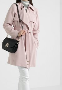 Heller rosa Trenchcoat mit einem taillierten Gürtel, großen Revers und Seitentaschen, kombiniert mit einer schwarzen Handtasche mit goldenem Logo. Weiße Hosen sind sichtbar.