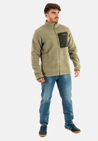 Giacca in fleece di un verde oliva chiaro con una tasca sul petto con zip. Presenta un colletto alto e un tessuto texturizzato. Abbinata a jeans blu e scarpe da ginnastica scure.