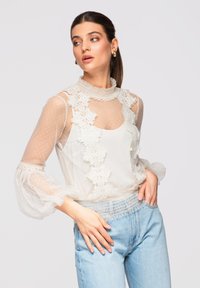 Femme en blouse blanche en dentelle transparente avec des poignets à volants et un jean taille haute bleu clair, regardant sur le côté avec la main dans la poche.