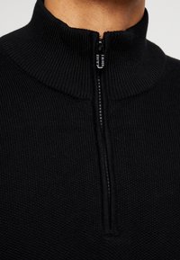 Pullover nero in maglia con mezzo zip, caratterizzato da un collare alto, tessuto testurizzato e una zip frontale con un tira-cerniera circolare.