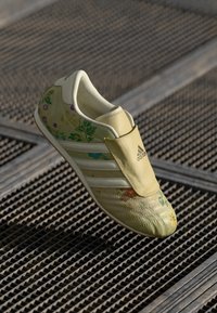 Světle žlutá sportovní bota s květinovými vzory, bílými pruhy a polstrovaným designem. Disponuje stylem na zvolením a strukturovanou podešví.