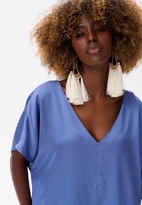 Luźna, niebieska bluzka z dekoltem w serek, krótkimi rękawami i gładką teksturą. Posiada duże białe kolczyki z frędzlami z złotymi akcentami.