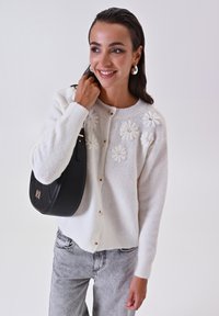 Cardigan bianco lavorato a maglia con applicazioni floreali, chiusura frontale con bottoni, colletto rotondo e polsini a coste. Abbinato a una borsa a spalla in pelle nera.