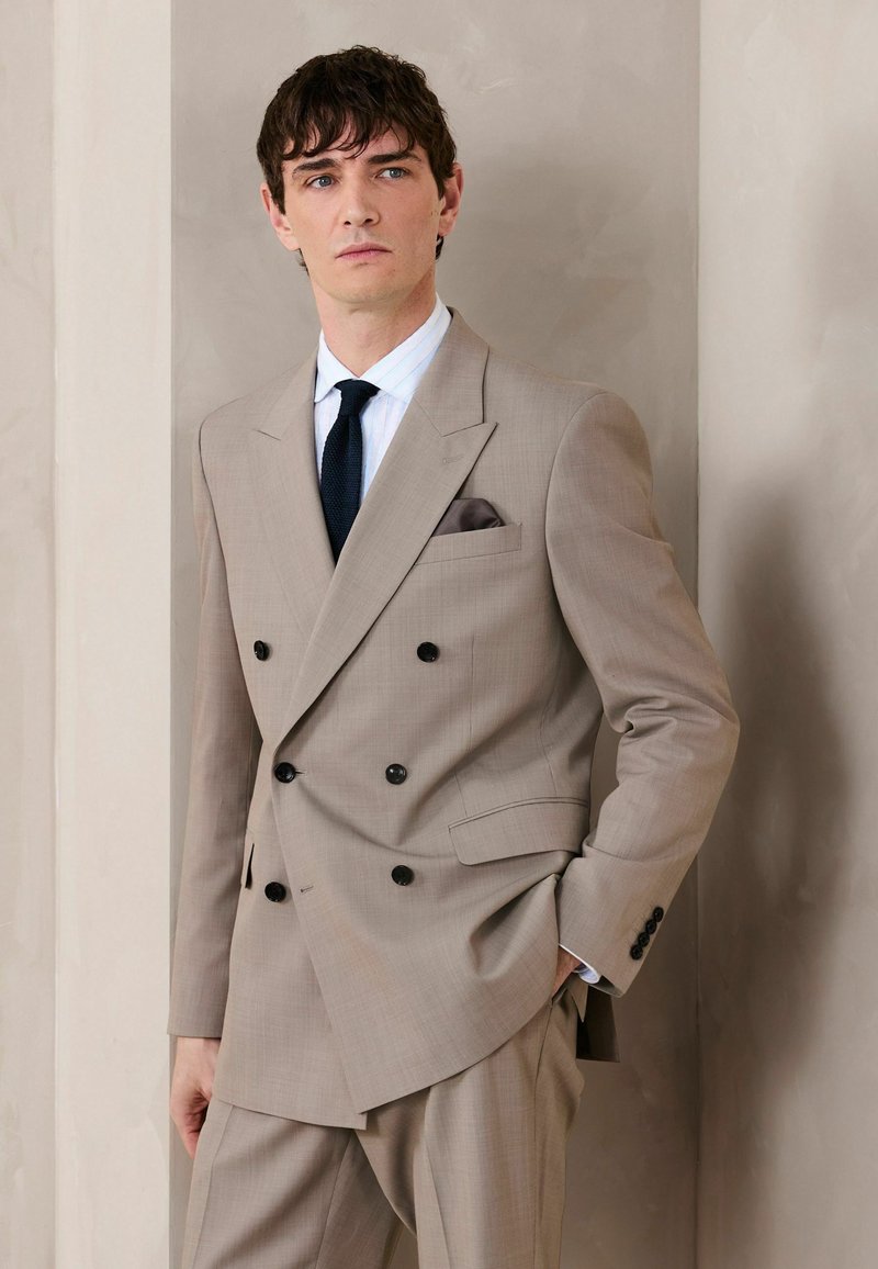 Homme portant un costume croisé beige avec des boutons noirs, une chemise blanche, une cravate noire et un mouchoir de poche marron, debout contre un mur neutre.