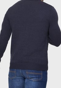 Dunkelblauer Strickpullover mit geripptem Ausschnitt und Bündchen. Verfügt über eine strukturierte Oberfläche und einen lässigen Schnitt. Getragen mit blauen Jeans.