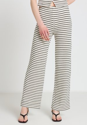 Frau trägt schwarz-weiß horizontal gestreifte, weit geschnittene Hose, schwarze offene Sandalen und ein passendes ärmelloses Top.
