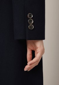 Morski wełniany płaszcz z rękawem ozdobionym trzema okrągłymi guzikami o chropowatej powierzchni. Dłoń znajduje się obok mankietu, ujawniając naturalne paznokcie.