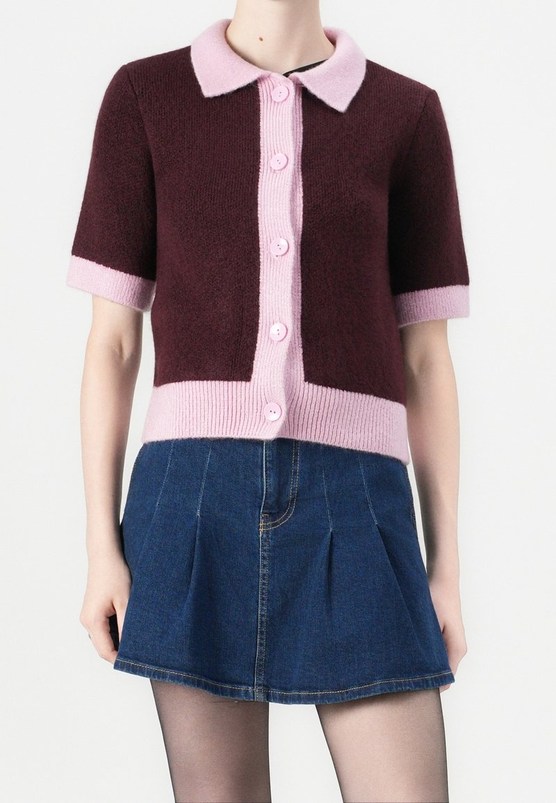 Femme portant un cardigan bordeaux à manches courtes avec col et bordure roses, boutonné sur le devant, associé à une mini-jupe plissée en denim bleu.