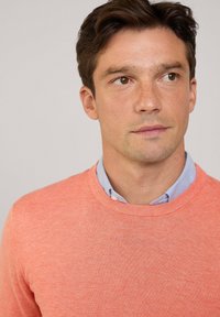 Jasnoróżowy sweter z teksturowanego dzianinowego materiału, z okrągłym dekoltem. Nosi się go na jasno-niebieskiej koszuli z kołnierzykiem.