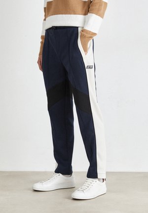 Marineblaue Jogginghosen mit schwarzen und cremefarbenen Seitenteilen, ausgestattet mit einem Kordelzugbund und seitlichen Taschen. Kombiniert mit weißen Sneakern.