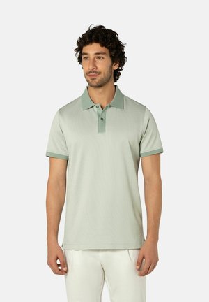 Mann trägt ein hellgrünes Poloshirt mit kurzen Ärmeln und weiße Hose, steht entspannt und blickt leicht zur Seite.