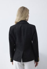 Kvinde med langt blondt hår iført en tekstureret sort blazer med guldknapper og hvide bukser, set bagfra mod en ensfarvet hvid baggrund.