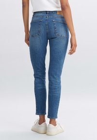 Žena v modrých úzkých džínách a bílých teniskách, stojící na světle šedém pozadí, pohled zezadu.