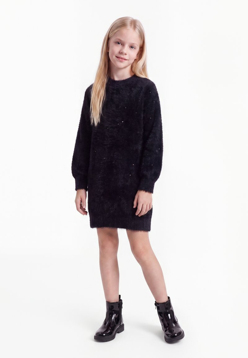 Fille blonde portant une robe noire à manches longues et duveteuse ainsi que des bottines noires brillantes, debout devant un fond blanc.