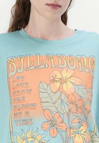 Camiseta de algodón color turquesa con un gráfico de estilo retro que presenta ilustraciones florales y el texto "BILLABONG" en naranja y azul.