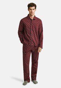 Man standing wearing maroon pajama set with paisley pattern and gray socks against plain white background.