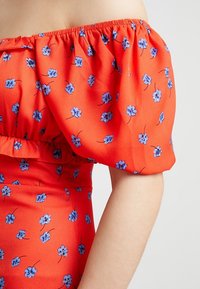 Červené květinové šaty s odhalenými rameny, zdobené nabíranými rukávy, vyrobené z lehké látky a s modrými květinovými vzory po celé ploše. Součástí je také volánkový lem.