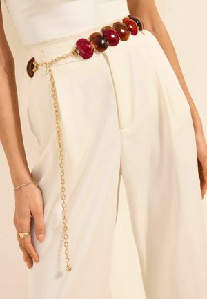 Baltas augsta vidukļa bikses ar dekoratīvu ķēdes jostu, kas rotāta ar lielām sarkanām un brūnām pērlēm un zelta toņa ķēdes akcentiem.