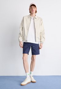 Lys beige bomberjakke, hvid t-shirt, marineblå shorts, lyse sneakers med gummisåler og pastelfarvet lyserød kasket. Simpelt design og afslappet pasform.