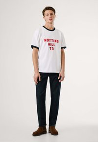 Λευκό βαμβακερό t-shirt με ναυτική περιφέρεια, που αναγράφει σε κόκκινο χρώμα "NOTTING HILL 73." Συνδυασμένο με σκούρα τζιν παντελόνια και καφέ μποτάκια έως τον αστράγαλο.