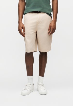 Man draagt beige shorts tot op de knie, witte sneakers, witte sokken en een groen shirt, staand tegen een effen lichte achtergrond.