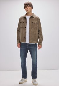 Hnědá kordová bunda se dvěma náprsními kapsami, zapínáním na knoflíky a rovný střih, spárovaná s modrými džíny a bílými teniskami.