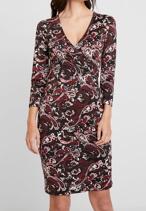 Frau trägt ein knielanges, langärmeliges Kleid mit einem Paisley-Muster in Rot, Weiß und Schwarz und einem V-Ausschnitt vor einem einfarbigen Hintergrund.