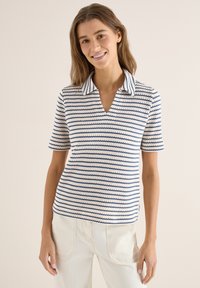 Femme souriante portant un polo en maille à manches courtes rayé blanc et bleu et un pantalon blanc cassé, sur un fond uni.