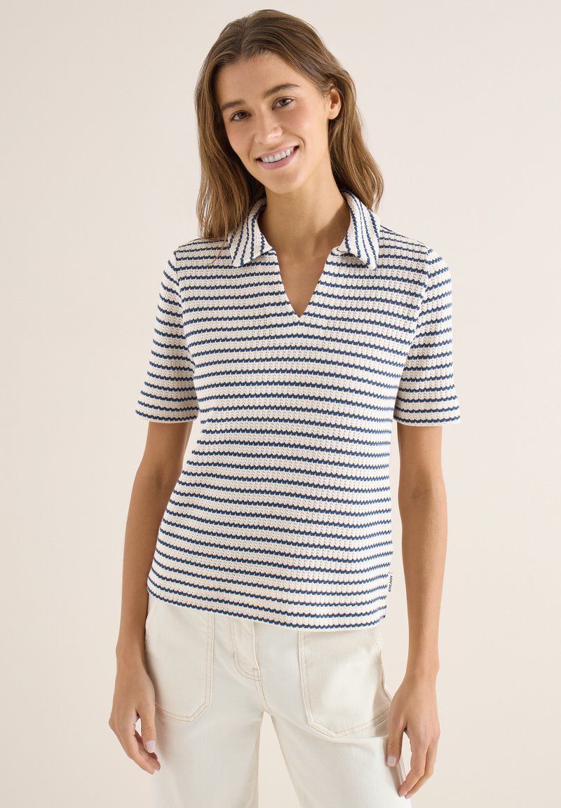 Femme souriante portant un polo en maille à manches courtes rayé blanc et bleu et un pantalon blanc cassé, sur un fond uni.