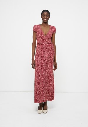 Γυναίκα που φοράει κόκκινο φόρεμα τύπου wrap με λευκές βούλες, κοντά μανίκια, ντεκολτέ σε σχήμα V και λευκά μυτερά παπούτσια με τακούνι, στέκεται μπροστά σε απλό φόντο.