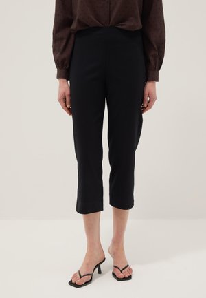 Frau, die schwarze kurze Hosen und schwarze Sandaletten mit Absatz trägt und vor einem einfachen hellen Hintergrund steht.