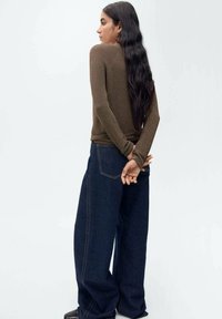 Maglia a maniche lunghe marrone in maglia con vita aderente, abbinata a jeans scuri a gamba larga. L'outfit presenta dettagli di cucitura sottili e una texture liscia.