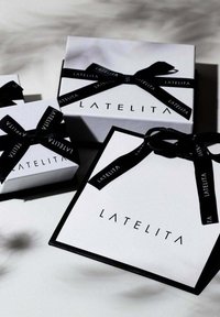 Tres cajas de regalo blancas de LATELITA, con cintas negras y logotipos. La caja más grande tiene un borde negro, mientras que la más pequeña es de forma cuadrada.