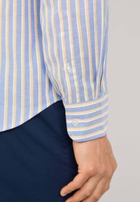 Camicia di cotone azzurro chiaro con righe gialle e bianche, polsini con bottoni e orlo curvato. Sono visibili dettagli di trama e motivo.