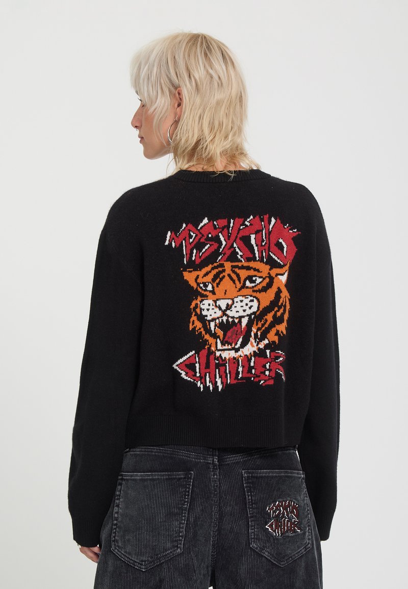 Personne aux cheveux blonds courts porte un pull noir avec un visage de tigre et un texte rouge "Psycho Chiller" dans le dos ainsi qu'un pantalon en velours côtelé foncé.
