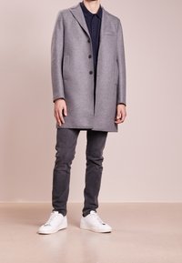 Šedý vlněný kabát s kusým límcem, několika knoflíky, bočními kapsami a hladkou texturou, doplněný tmavými džínami a bílými teniskami.