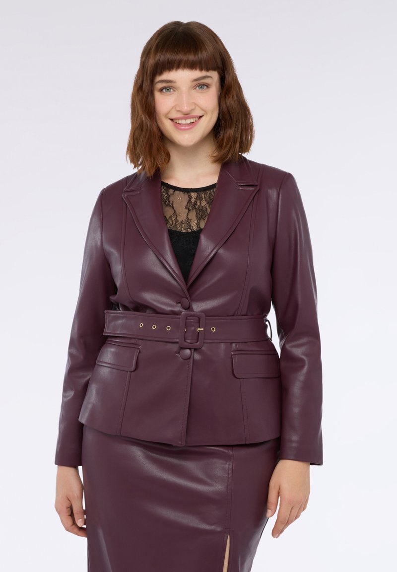 Śliwkowa marynarka z imitacji skóry z paskiem w talii, z klapami i dwiema przednimi kieszeniami, w połączeniu z czarnym koronkowym topem i pasującą spódnicą.