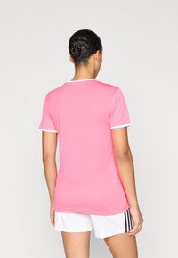 Ροζ αθλητική μπλούζα με κοντά μανίκια και λευκές λεπτομέρειες, κατασκευασμένη από λείο ύφασμα. Φορεμένη με λευκά σορτς που έχουν μαύρες ρίγες.