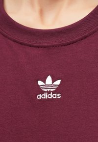 Bordó pamut pulóver bordázott kerek nyakkal és fehér hímzett Adidas logóval, három levélszerű díszítéssel a mellrészen.